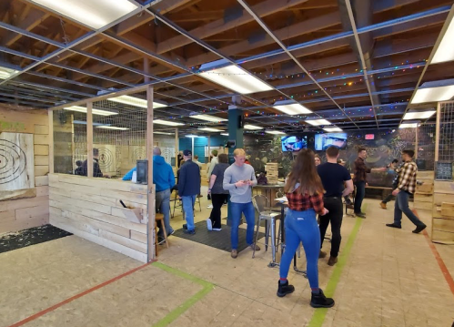 A lively indoor axe-throwing venue with people socializing and wooden targets in the background. Colorful lights adorn the space.