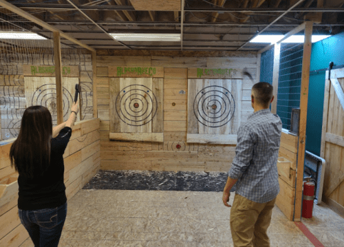Two people in an indoor axe-throwing venue, preparing to throw axes at wooden targets with painted bullseyes.