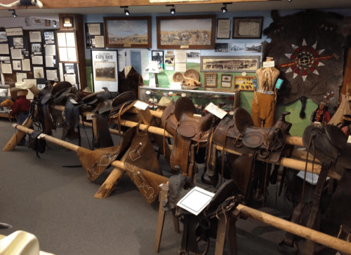 A display of various saddles and cowboy artifacts in a museum setting, with historical information on the walls.