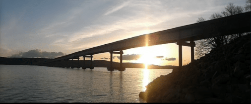 A silhouette of a bridge over water at sunset, with a colorful sky and distant hills in the background.