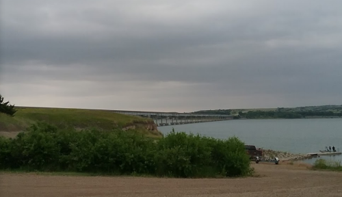 A bridge spans over a calm body of water, with a cloudy sky and green vegetation in the foreground.
