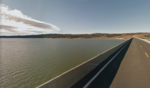 A wide view of a road alongside a calm lake, with hills in the background under a clear blue sky.
