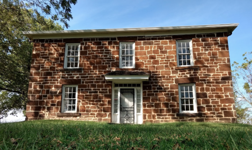 A two-story stone house with multiple windows, set against a clear blue sky and grassy foreground.
