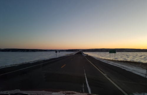 A long, empty road stretches into the horizon, flanked by frozen water under a colorful sunset sky.