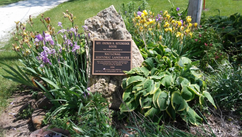 A stone marker for the Rev. George B. Hitchcock House, surrounded by colorful flowers and greenery.