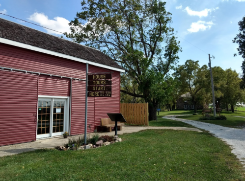 A pink barn with a sign for house tours, surrounded by greenery and a gravel path leading into the distance.