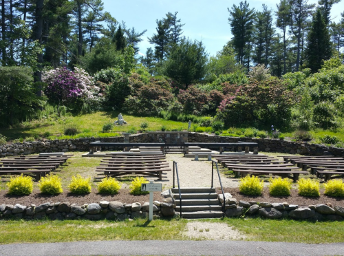 An outdoor amphitheater surrounded by lush greenery and blooming flowers, with stone seating and a pathway.