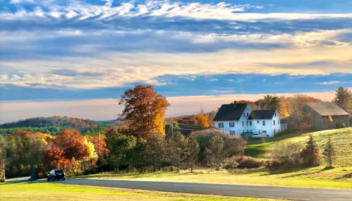 A scenic autumn landscape featuring a white house, colorful trees, and a clear sky with clouds.