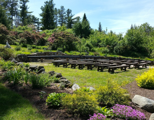 An outdoor amphitheater with wooden benches surrounded by greenery and colorful flowers under a clear blue sky.