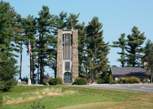 A stone tower surrounded by tall trees, with a small building nearby and an American flag in the foreground.