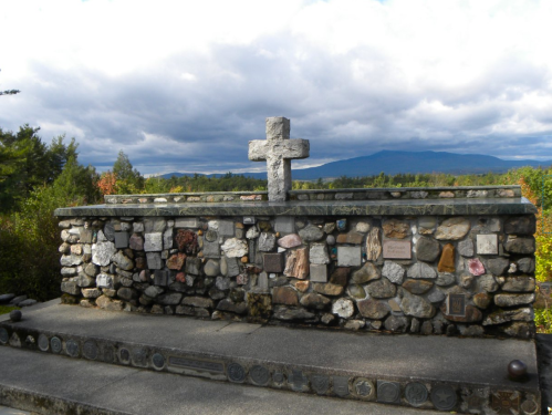 A stone memorial with a cross, surrounded by plaques, set against a backdrop of mountains and cloudy skies.