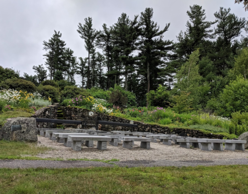A stone amphitheater surrounded by lush greenery and flowers, set against a backdrop of tall trees under a cloudy sky.