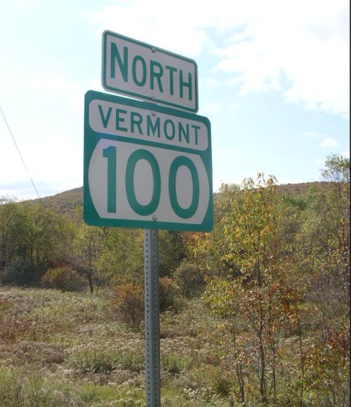 Sign indicating "North Vermont 100" with a scenic background of trees and hills under a partly cloudy sky.