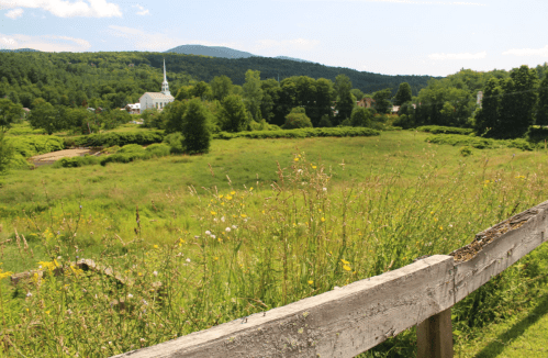 A scenic view of a green field with wildflowers, a wooden fence, and a church in the background against a mountain backdrop.