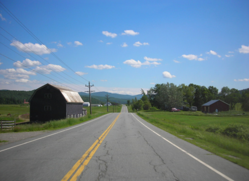 A rural road stretches ahead, flanked by green fields and a barn under a blue sky with scattered clouds.