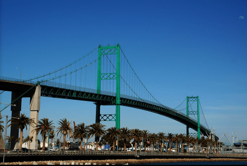 A green suspension bridge spans over a waterfront lined with palm trees under a clear blue sky.