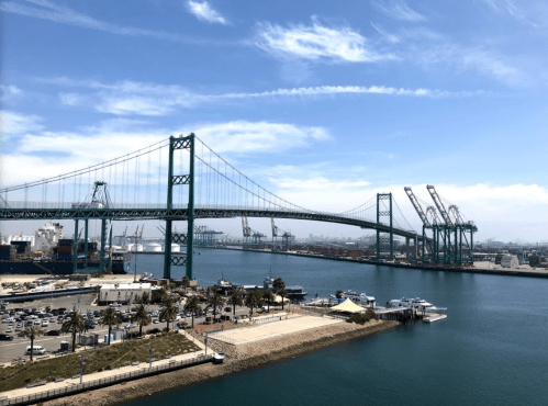 A view of a green bridge over water, with ships and cranes in the background under a clear blue sky.