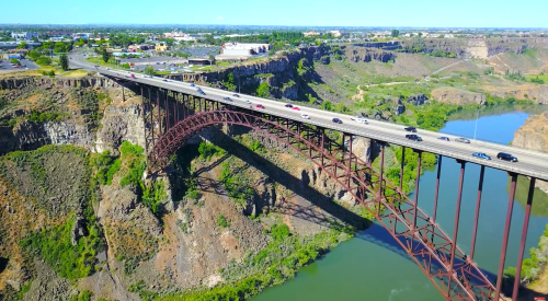 Aerial view of a large bridge spanning a canyon, with cars traveling and a river flowing below. Lush greenery surrounds the area.