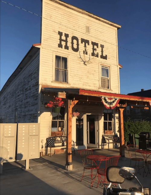 A rustic hotel building with a wooden porch, red chairs, and flower pots, set against a clear blue sky.