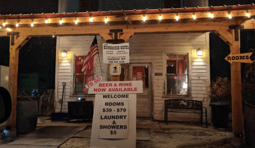 A cozy hotel entrance with a sign offering rooms, laundry, showers, and to-go beer and wine, decorated with lights.