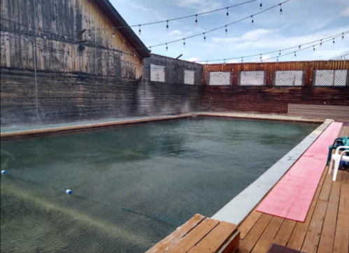 A serene outdoor pool surrounded by wooden walls, with mist rising and string lights overhead.