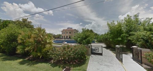 A large, elegant house with a pool, surrounded by lush greenery and colorful flowers, viewed from a gated entrance.