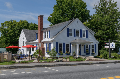 A charming white house with blue shutters, featuring a porch and outdoor seating under red umbrellas, surrounded by greenery.