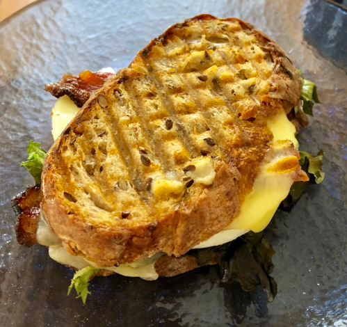 A toasted sandwich with crispy edges, filled with greens, cheese, and bacon, served on a glass plate.