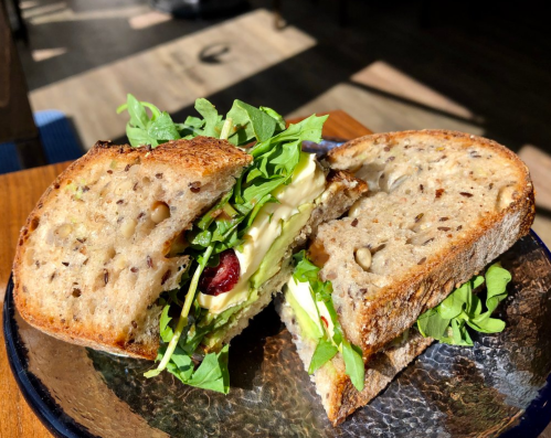 A close-up of a sliced sandwich on a plate, filled with greens, creamy spread, and seeds in the bread.