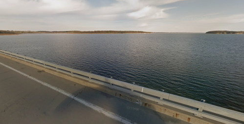A serene view of a calm lake with a distant shoreline, seen from a bridge under a partly cloudy sky.