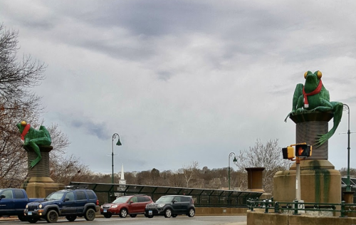 Two large green frog sculptures wearing red scarves sit atop pillars by a road, with cloudy skies in the background.