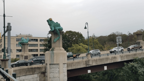 Two frog sculptures sit atop pillars on a bridge, with cars passing below and trees in the background.