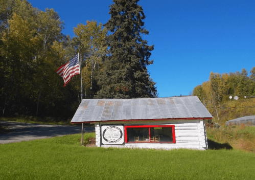 A small log cabin with a metal roof, featuring a sign and an American flag, surrounded by green grass and trees.