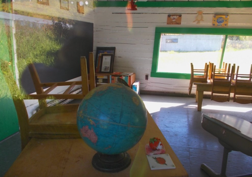 A rustic classroom with a globe on a desk, empty chairs stacked, and sunlight streaming through a window.