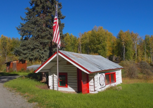 A small log cabin with a red door and a flag, surrounded by trees and greenery under a clear blue sky.