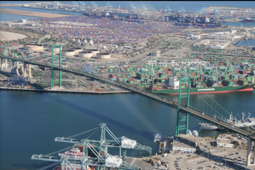 Aerial view of a busy port with shipping containers, a bridge, and cranes along the waterfront.