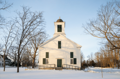 A white church with a bell tower, surrounded by snow-covered ground and bare trees under a clear blue sky.
