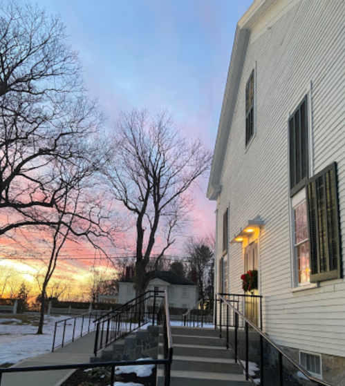 A white building with a staircase, surrounded by bare trees, against a colorful sunset sky. Snow is visible on the ground.