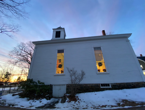 A white building with large windows, surrounded by snow, under a colorful sunset sky.
