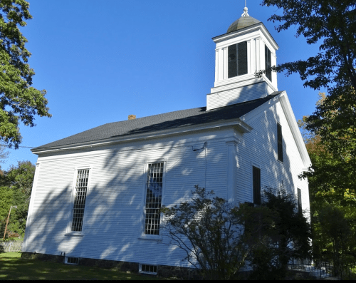 A white church building with a steeple, surrounded by trees and a clear blue sky.