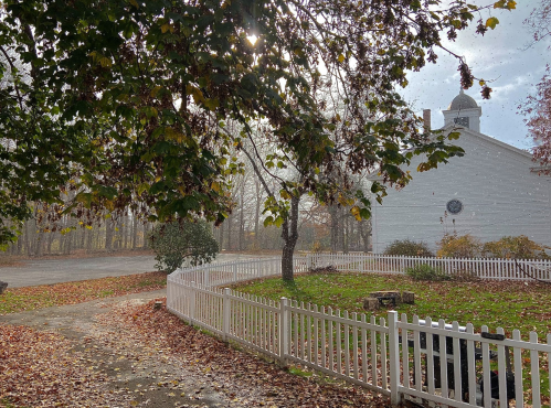 A serene scene of a white building surrounded by autumn leaves and a white picket fence, with sunlight filtering through trees.