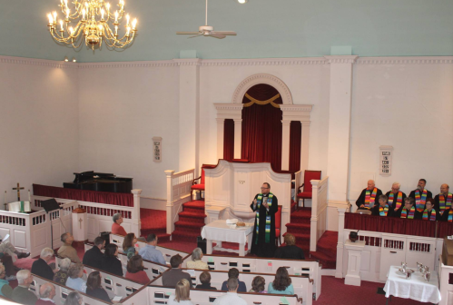 A pastor speaks at the front of a church, with congregants seated and choir members in colorful stoles behind him.