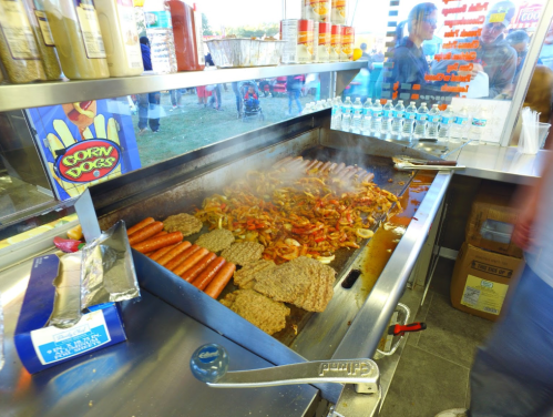A grill filled with sizzling hot dogs, burgers, and sautéed peppers, with people in the background at a food stand.
