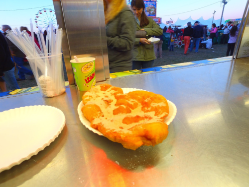 A plate of funnel cake dusted with powdered sugar, with a fair atmosphere in the background.