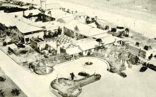 Aerial view of a historic building complex with gardens and parked cars, near a beach or coastal area.