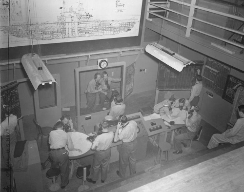 A historical control room with personnel working at desks, wearing headsets, and monitoring large maps and screens.