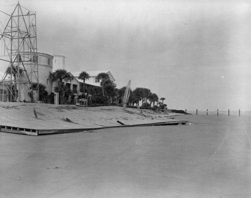 Black and white photo of a beachside building with palm trees and a construction structure in the foreground.