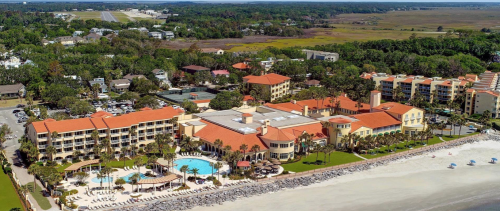 Aerial view of a coastal resort with a pool, palm trees, and nearby beach, surrounded by greenery and a runway in the distance.