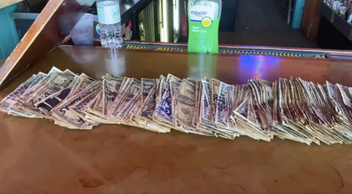 A long row of cash bills laid out on a wooden bar counter, with a bottle of sunscreen and water in the background.