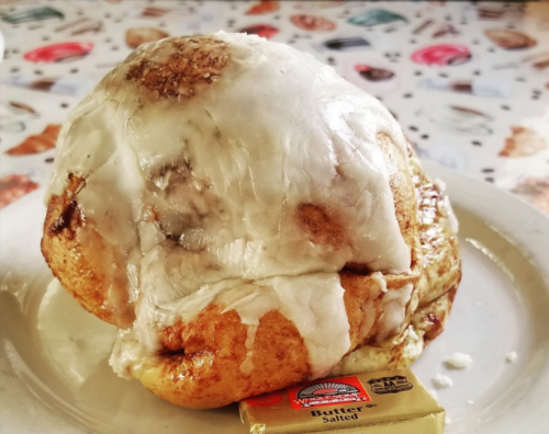 A large, frosted cinnamon roll sits on a plate next to a pat of salted butter.
